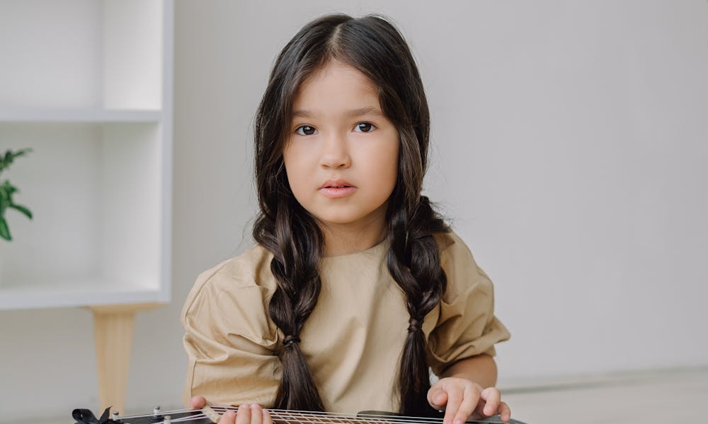 A young girl in a brown dress sits on a rug, playing a ukulele in a cozy room. .pexels