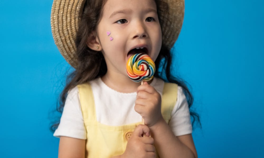 Charming girl in a straw hat and yellow sundress enjoying a colorful lollipop. .pexels