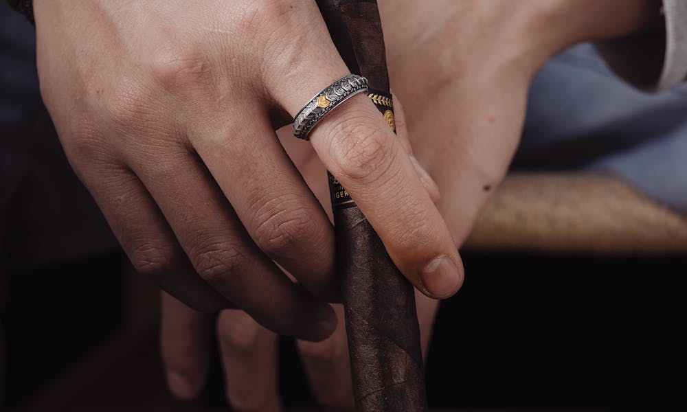 A close-up of a person's hands with rings, holding a cigar over a leather journal, symbolizing luxury and relaxation. .pexels