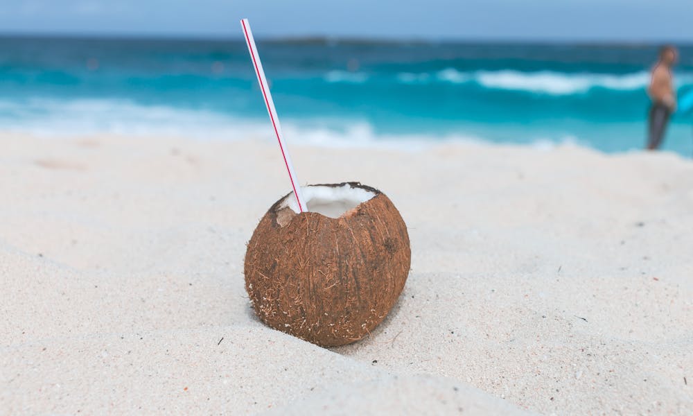 A coconut with a straw sits on a sandy beach, overlooking the clear blue ocean. Perfect tropical refreshment. .pexels