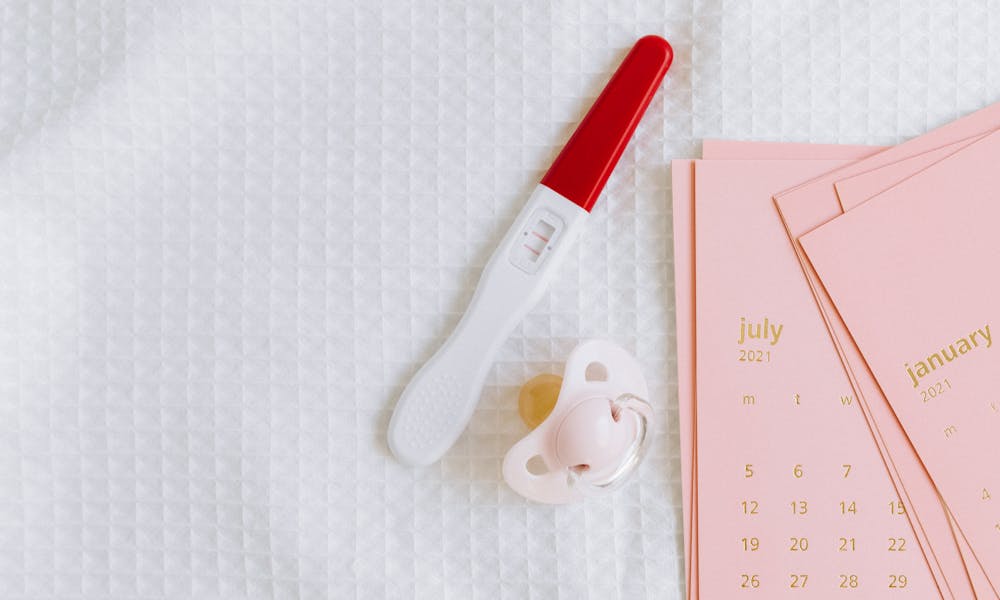 A pregnancy test with a positive result next to a baby pacifier and calendar on a textured surface. .pexels