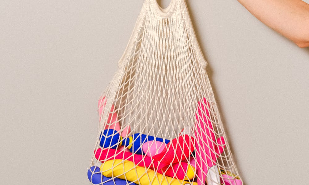 A mesh bag filled with colorful silicone sex toys held by an adult's hand against a neutral background. .pexels