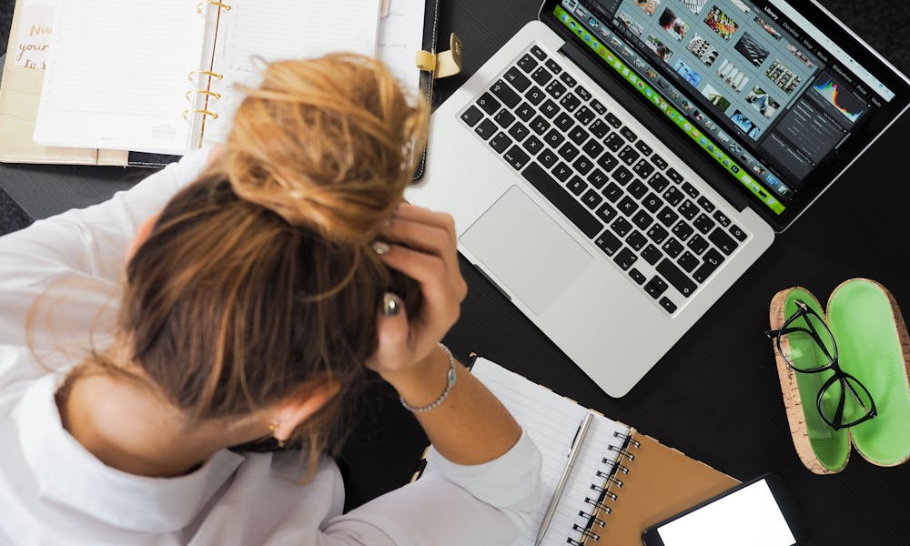 Overhead view of a stressed woman working at a desk with a laptop, phone, and notebooks. .pexels