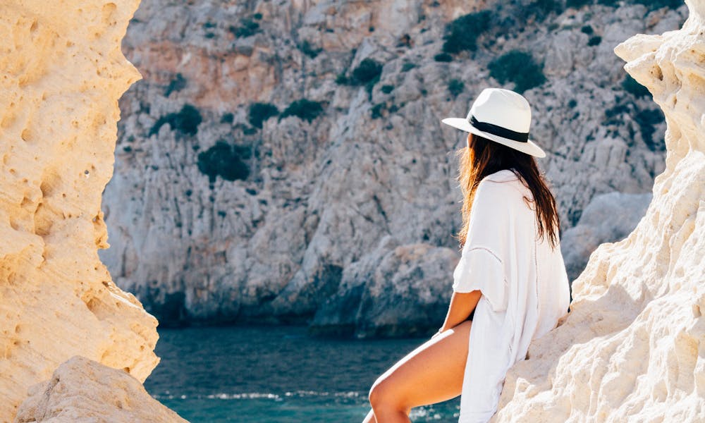 A woman in a hat sits on a rocky beach in summer, enjoying the sea view. .pexels