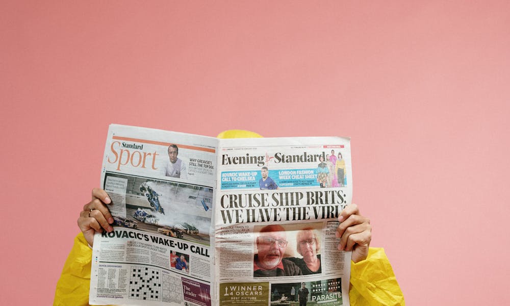 Individual in protective suit reads newspaper with pandemic headline against pink backdrop, symbolizing COVID-19 era. .pexels