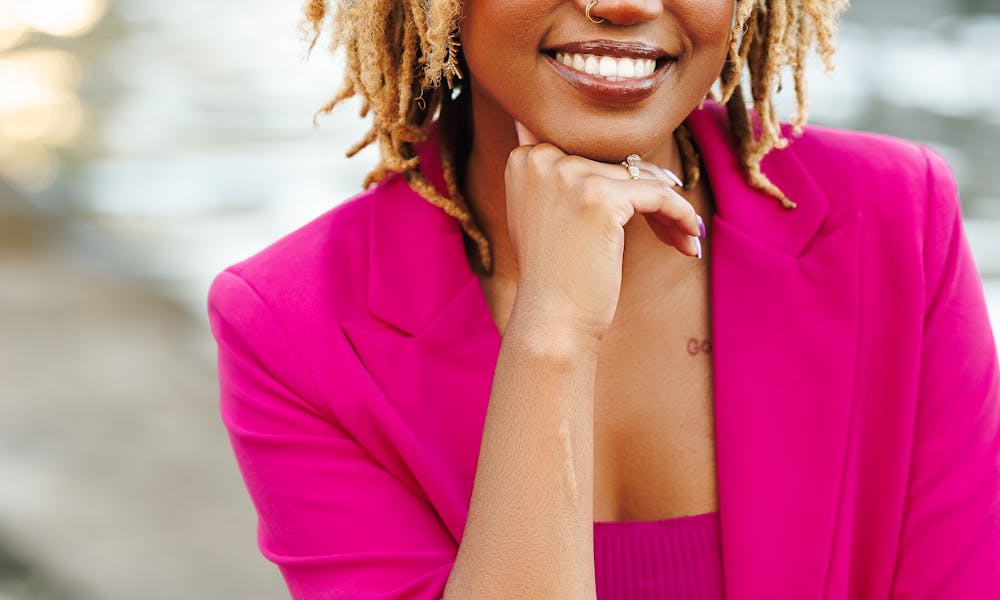 Confident woman in a pink suit smiling by the water, exuding warmth and positivity. .pexels