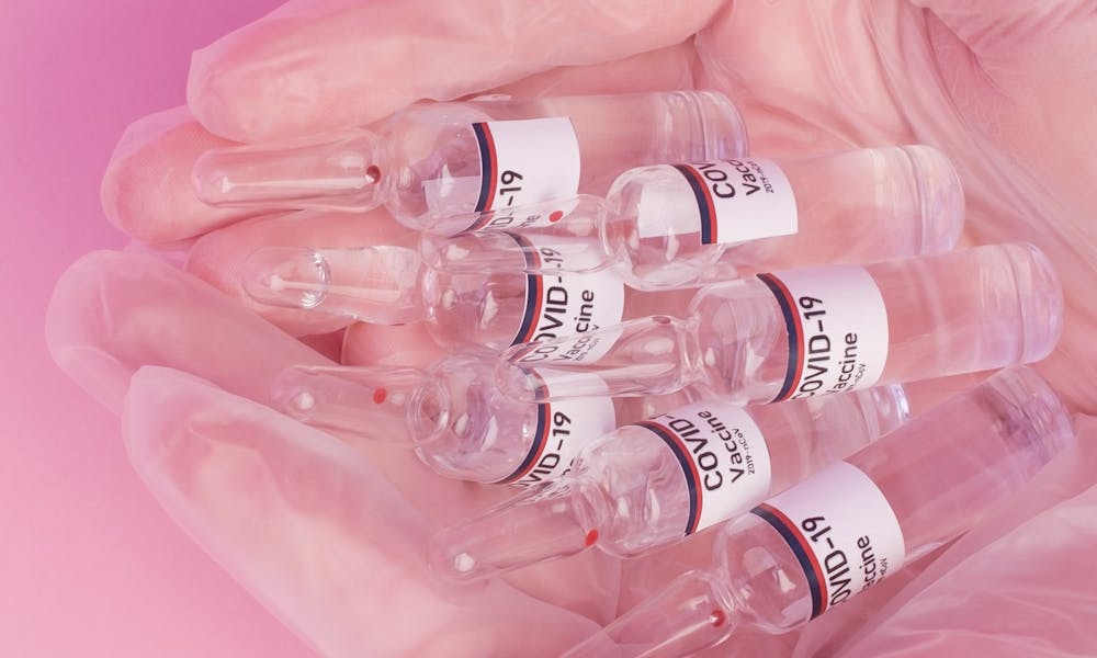 Gloved hands holding COVID-19 vaccine ampoules against a pink studio backdrop, symbolizing protection and healthcare. .pexels