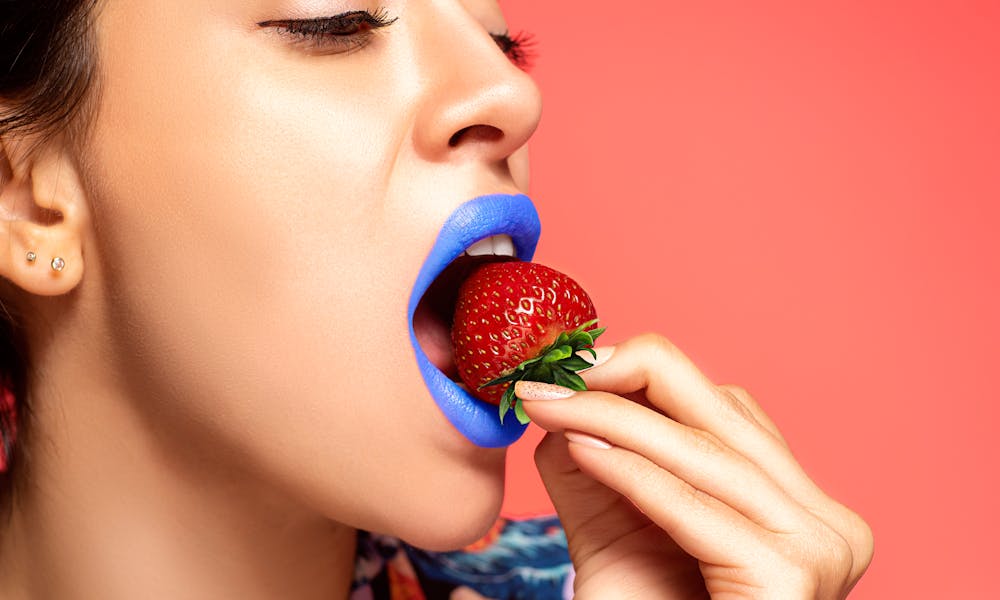 Colorful close-up of a woman with blue lipstick eating a ripe strawberry. .pexels