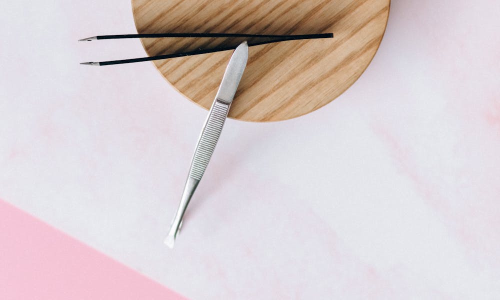 A high-angle shot of tweezers placed on a wooden coaster against a pastel pink background. .pexels