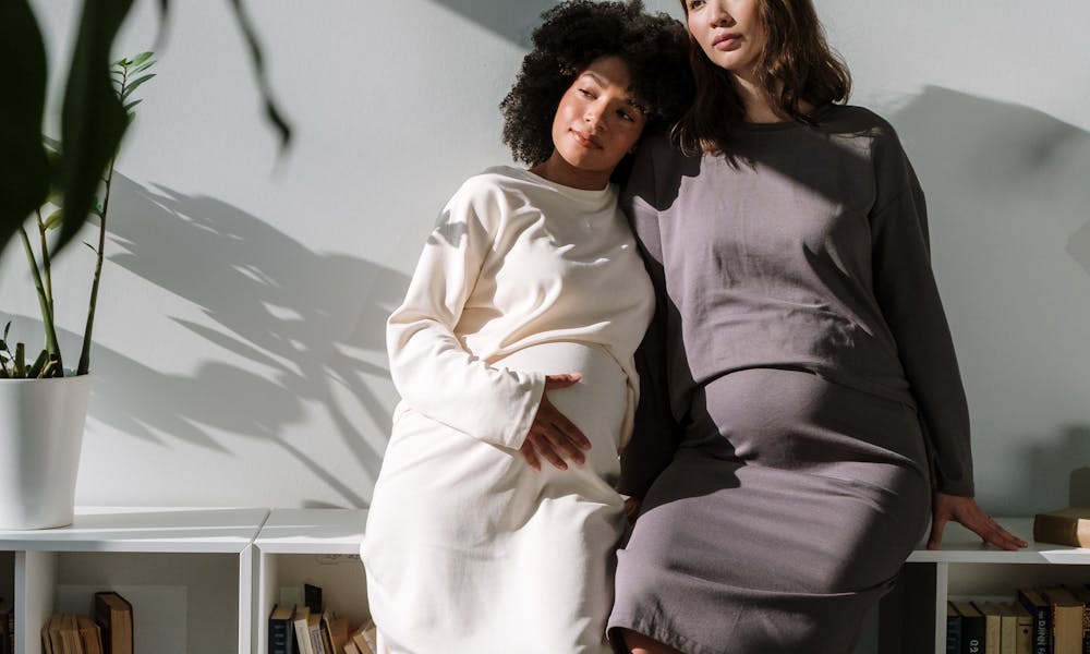 Two expectant mothers in stylish dresses, posing with a bookshelf backdrop, embodying maternity elegance. .pexels