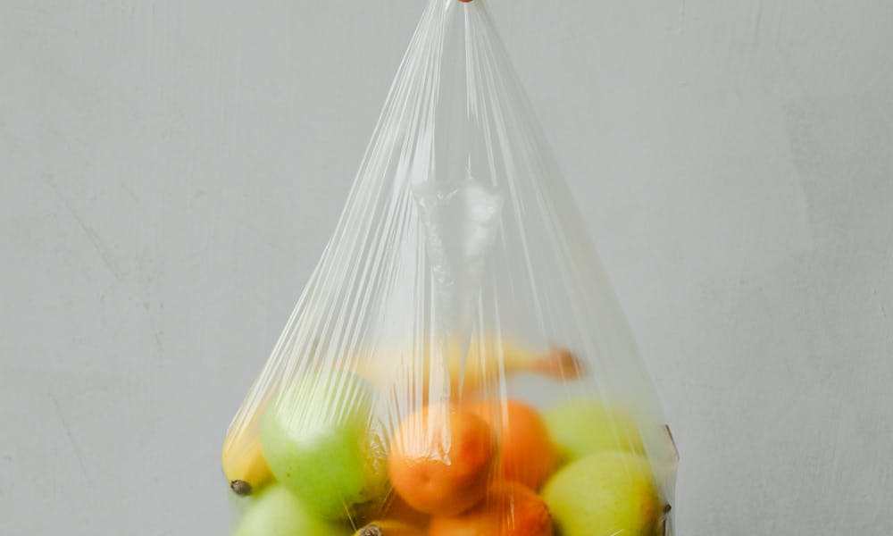 A hand holding a transparent plastic bag full of fresh citrus fruits against a neutral background. .pexels