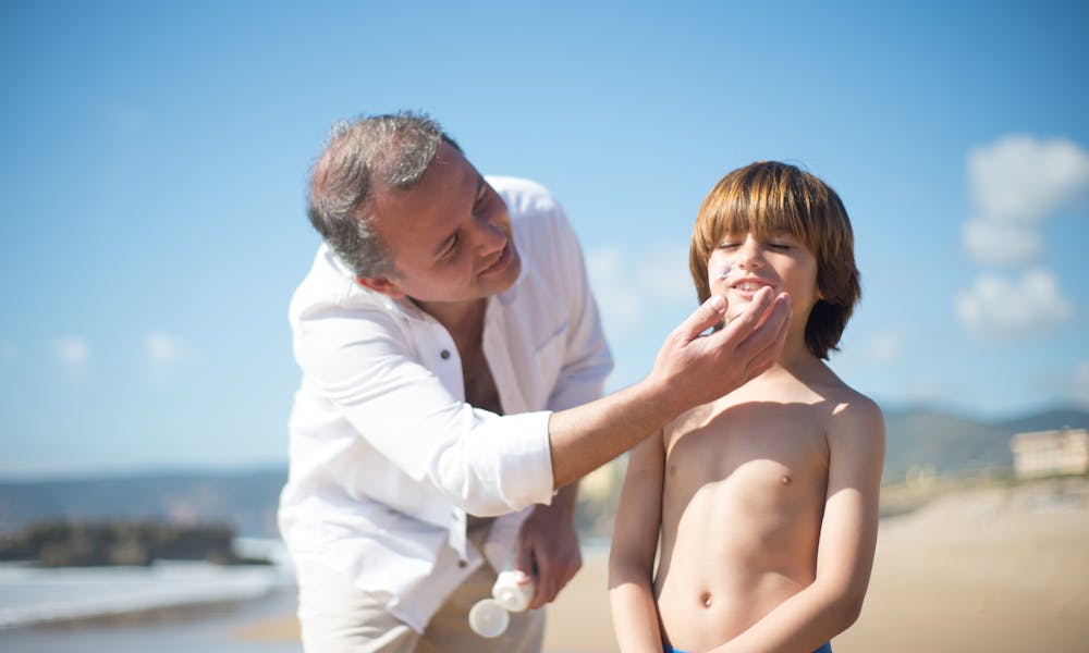 A father applies sunscreen to his smiling son on a sunny beach day. .pexels