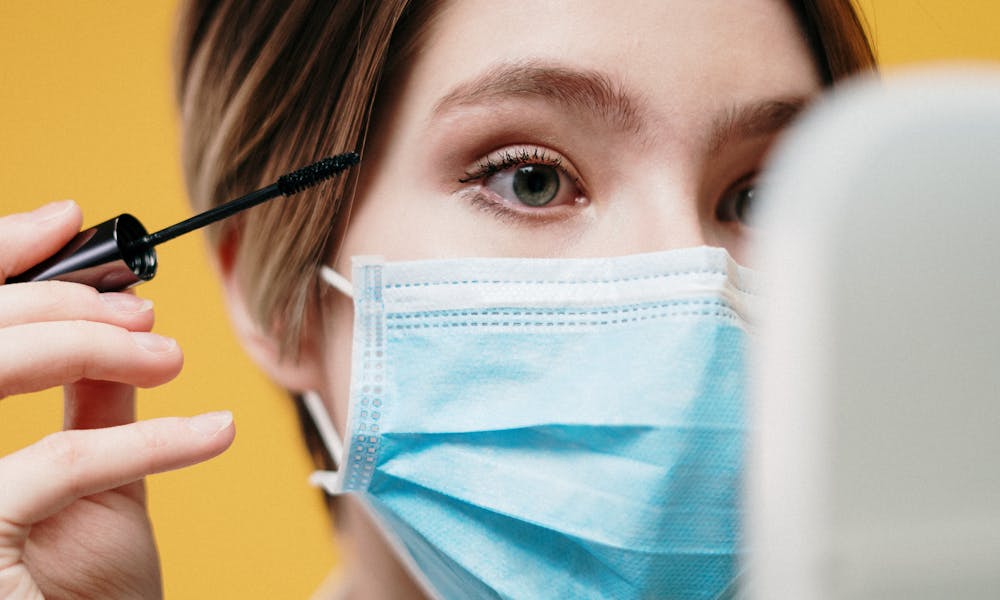 Woman applying mascara with a surgical mask on, reflecting pandemic beauty routines. .pexels