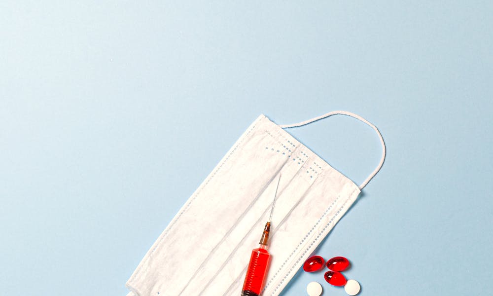 Flat lay of face mask, syringe, and pills on a blue surface, symbolizing healthcare themes. .pexels