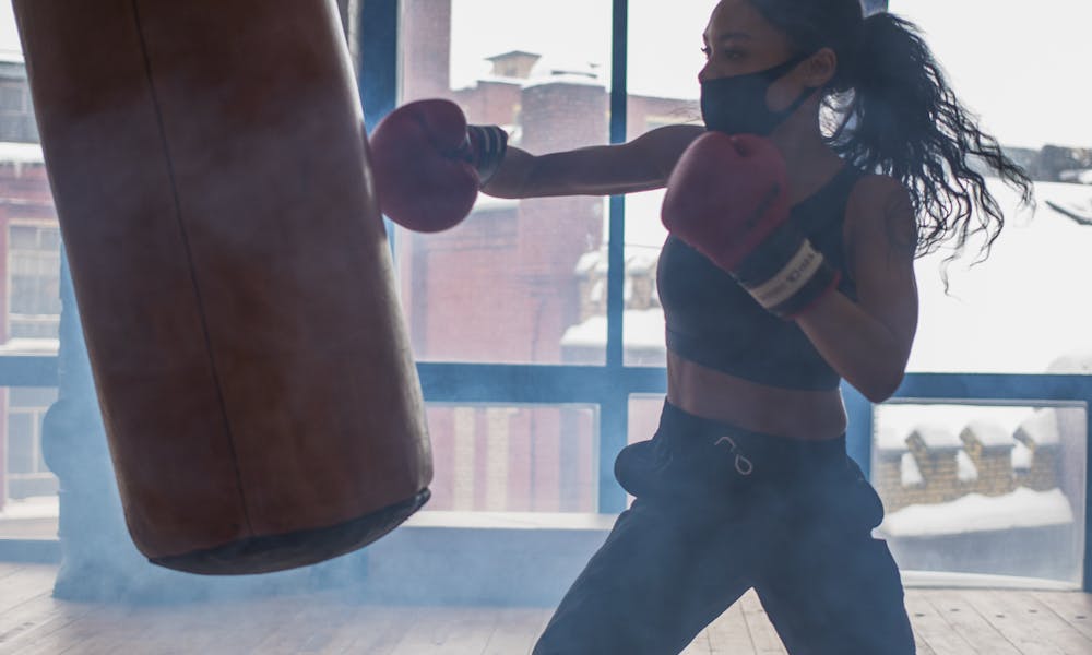 A woman boxer in a mask trains with a punching bag indoors, exemplifying focus and strength. .pexels