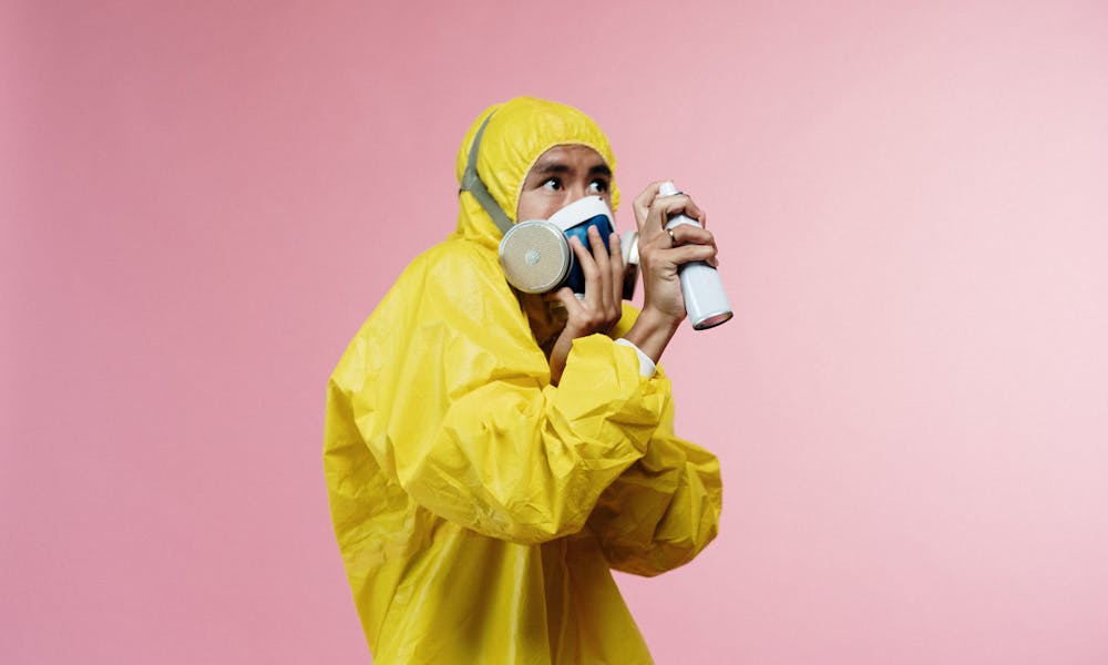 A person in a yellow hazmat suit holding a spray can against a pink background. .pexels