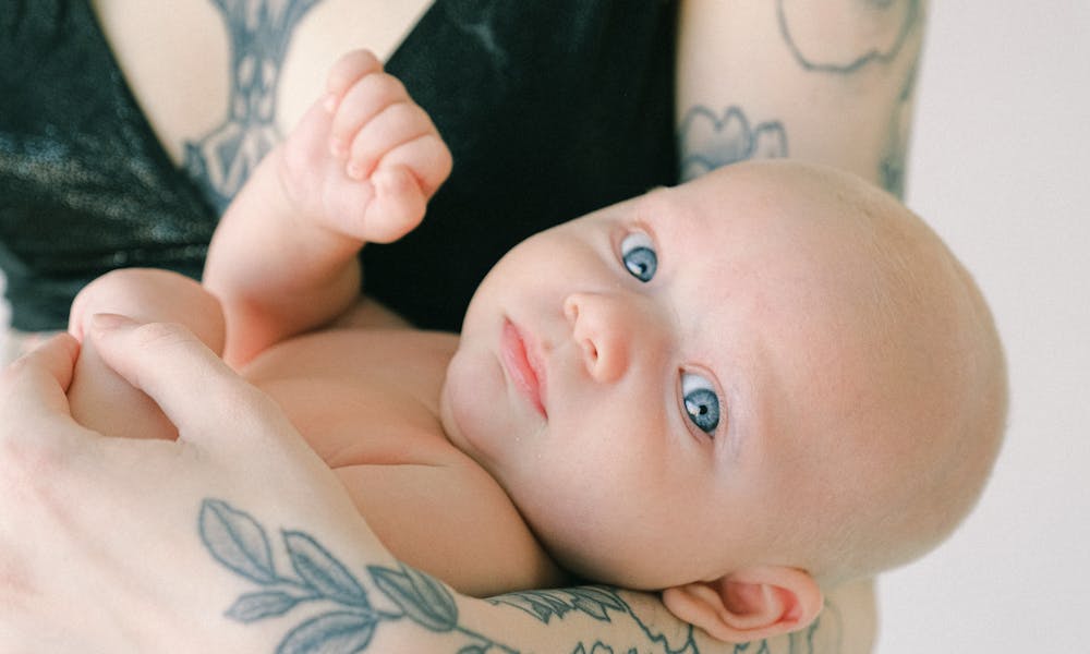 A beautiful baby with blue eyes held gently by tattooed arms, showcasing love and innocence. .pexels