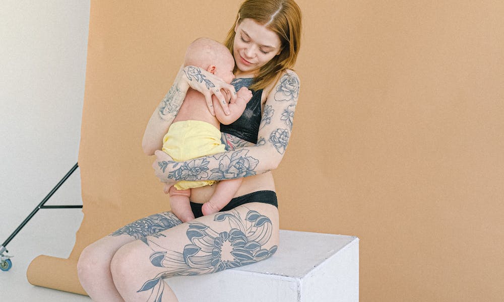 A tattooed mother lovingly embraces her infant in a serene studio setting. .pexels