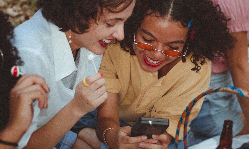 A group of happy young women enjoying a sunny picnic outdoors, engaging with a smartphone and laughing. .pexels