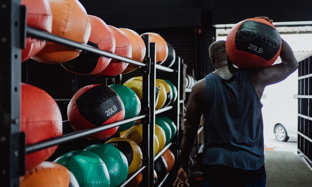 Back view of a man carrying a 20LB medicine ball in a gym with colorful balls on racks. .pexels