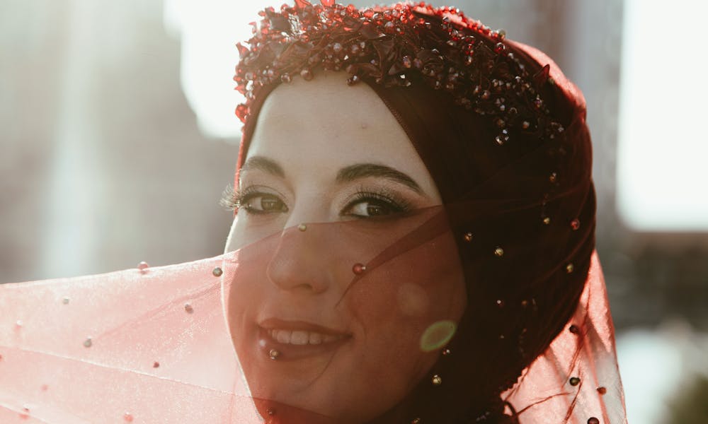 Close-up of a woman in a red veil adorned with beads, smiling softly in the sunlight. .pexels