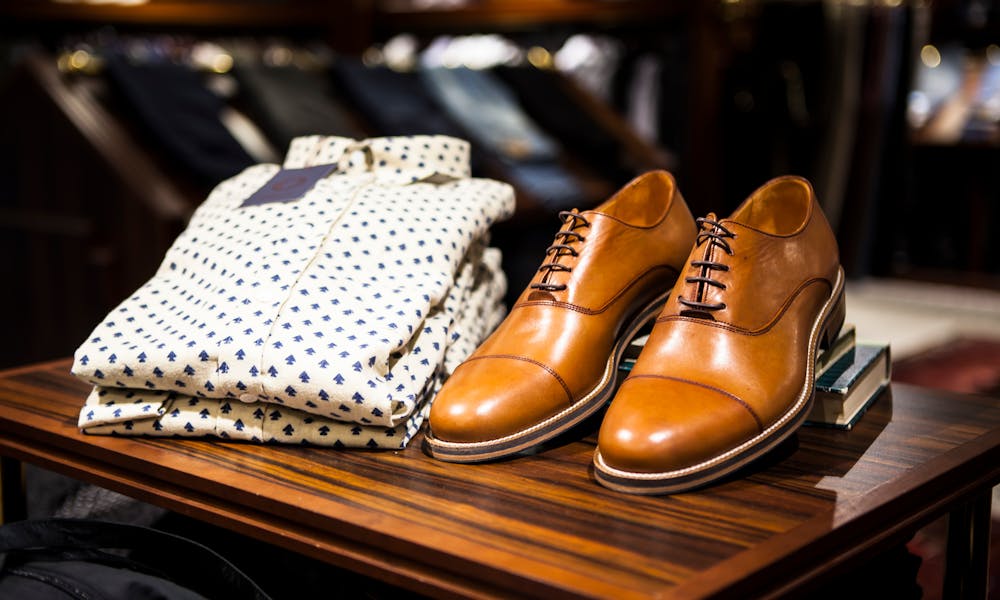 Elegant brown leather shoes and printed shirt displayed on a wooden table in a stylish clothing store. .pexels