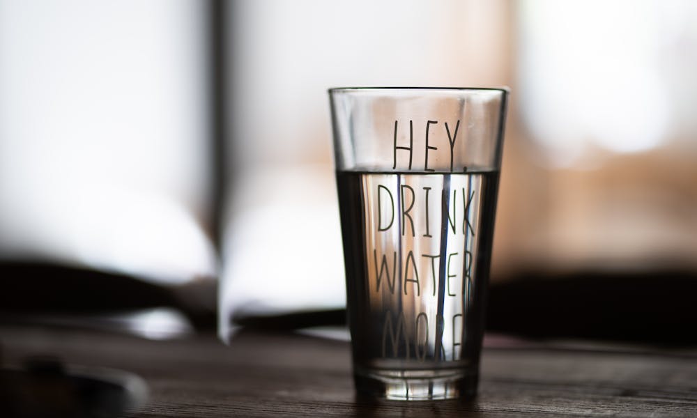 Close-up of a water glass with motivational text 'Hey Drink Water More' in a blurred indoor setting. .pexels