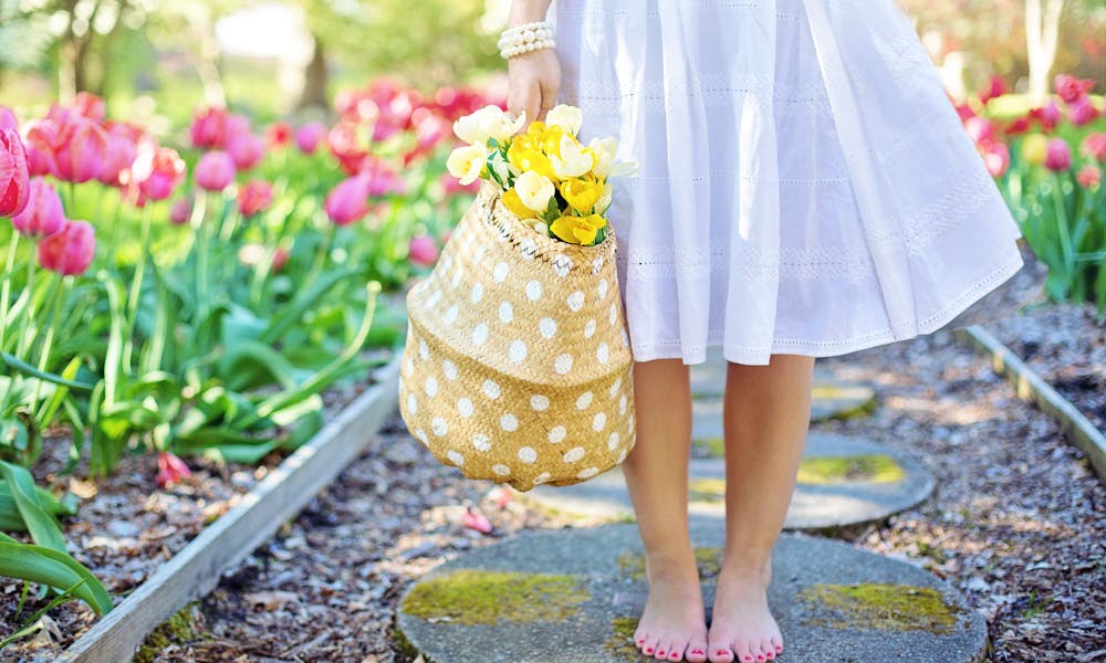 A woman in a white dress carries a basket of yellow flowers while walking barefoot in a tulip garden. .pexels