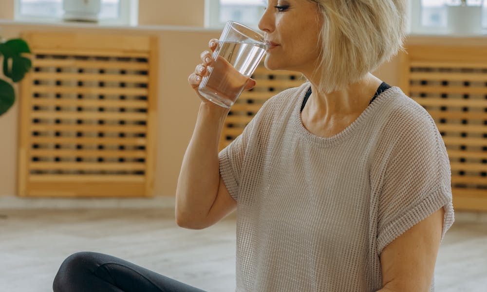 Senior woman in black leggings drinking water while seated on a yoga mat indoors. .pexels