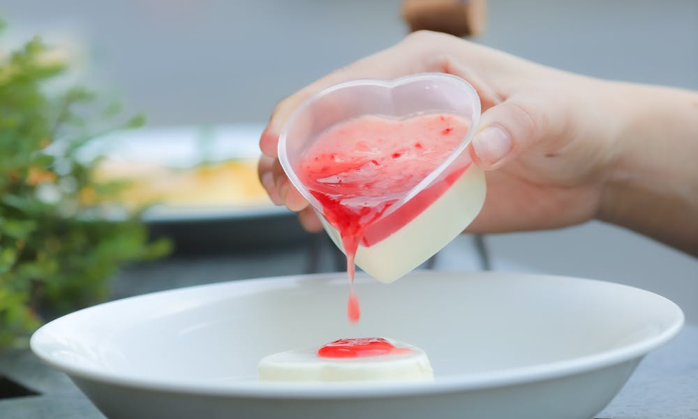 A person pours red syrup onto a heart-shaped dessert in an elegant white bowl. .pexels