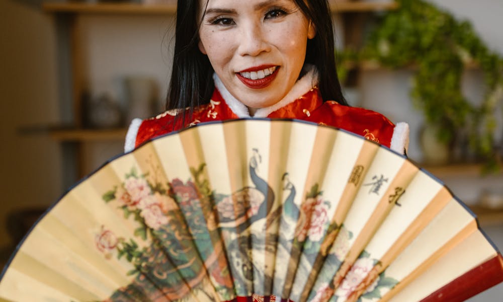 An elegant woman in red traditional attire holds a beautiful folding fan indoors. .pexels